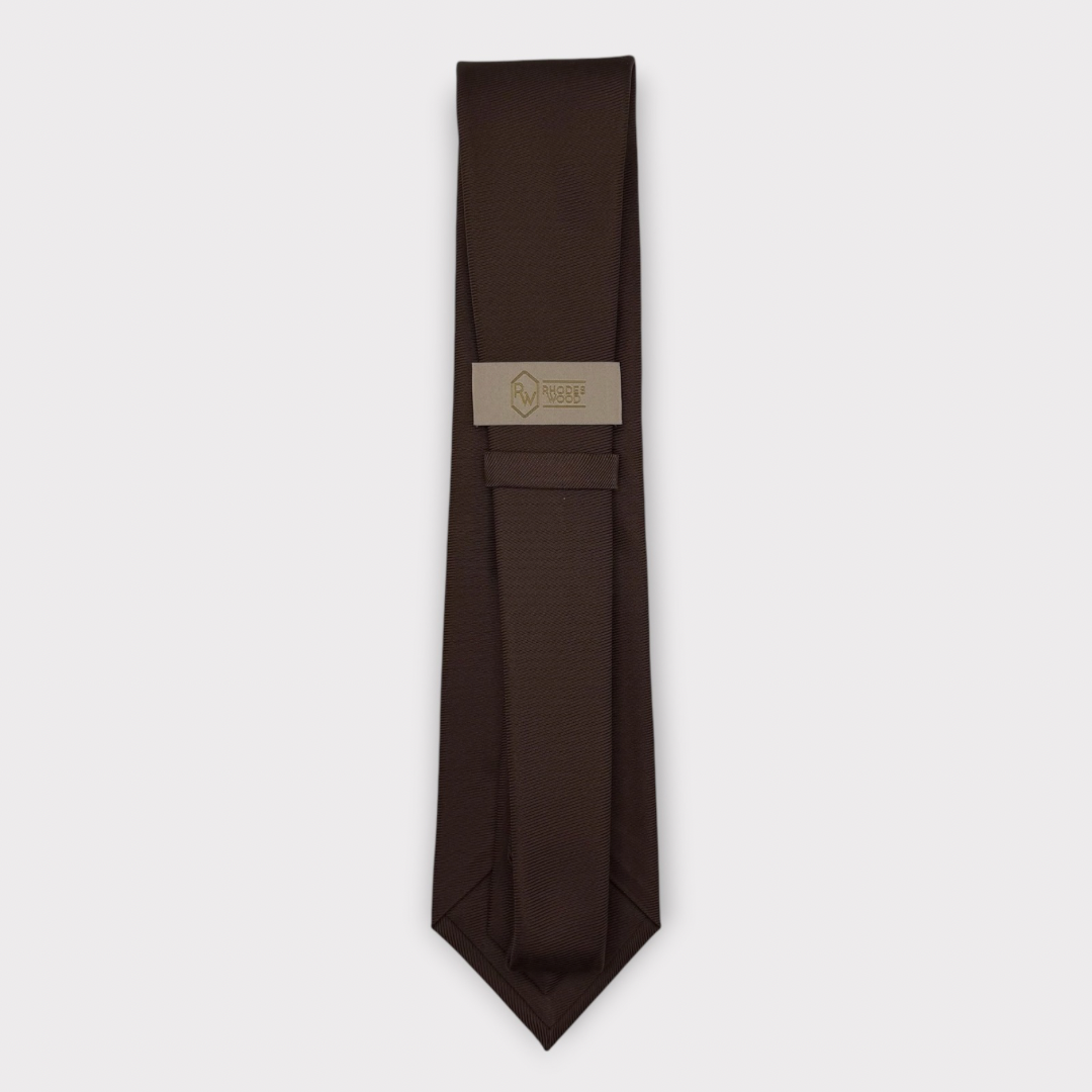 A dark brown necktie with a subtle textured pattern, laid flat on a light gray background. A beige paper label with a gold logo is attached near the collar area.