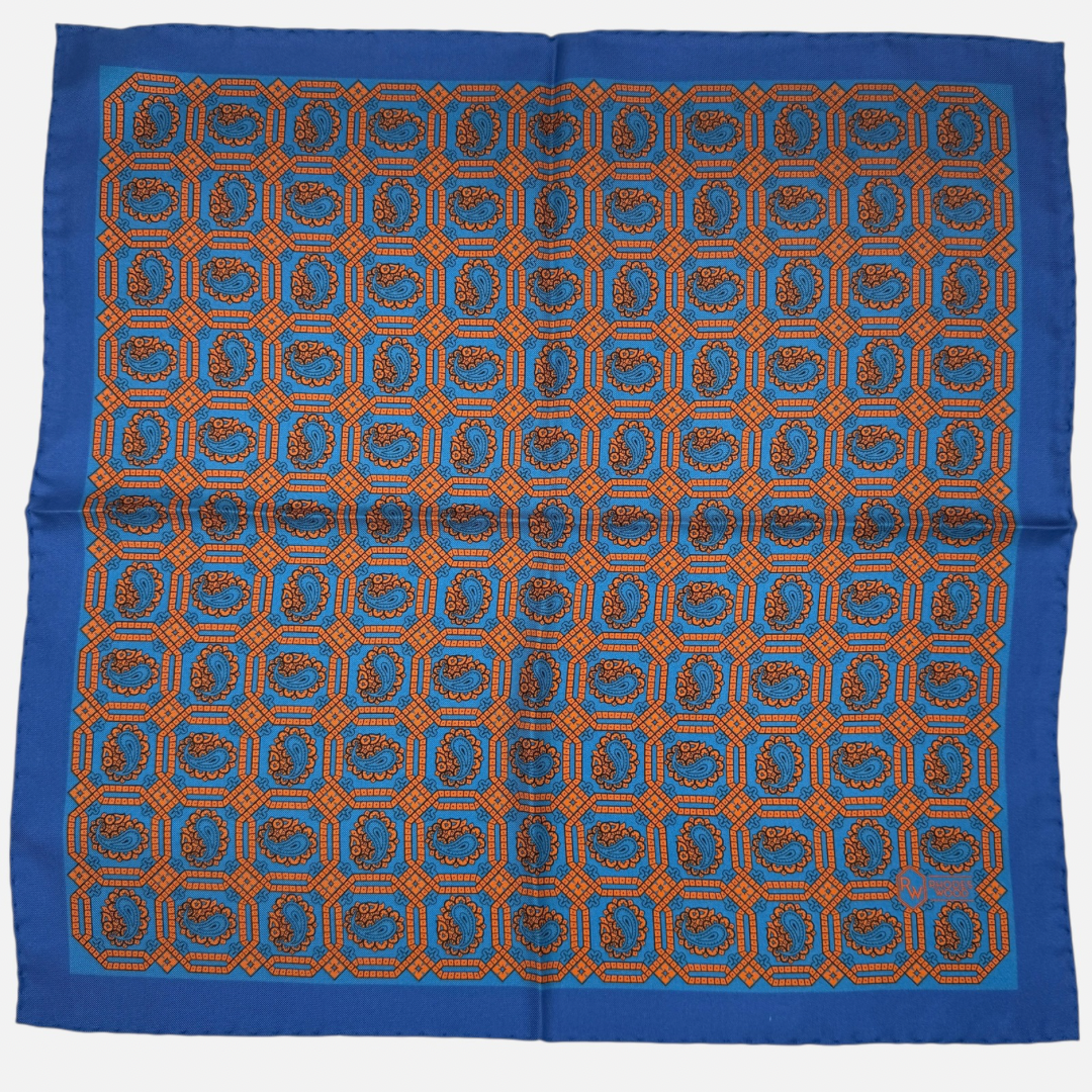 A blue silk pocket square with an orange octagon paisley repeating across the silk. 