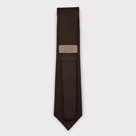 A dark brown necktie with a subtle textured pattern, laid flat on a light gray background. A beige paper label with a gold logo is attached near the collar area.