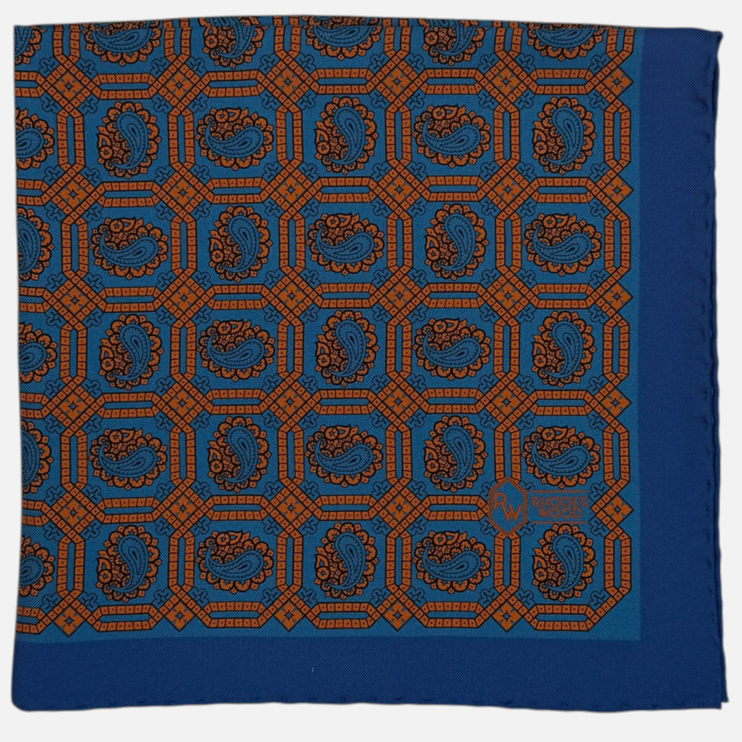 A blue silk pocket square with an orange octagon paisley repeating across the silk. 