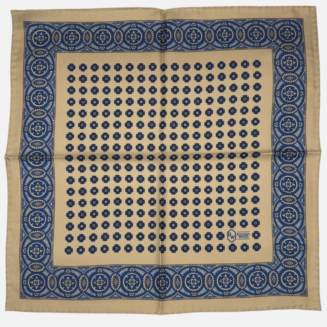 A camel 100% silk pocket square with navy blue medallions repeating across the silk .