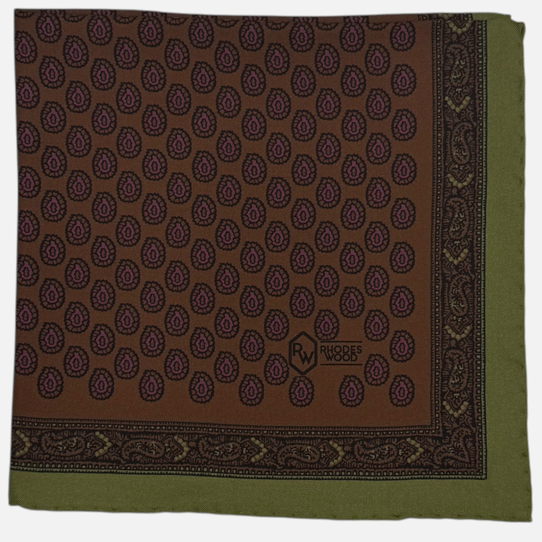 A brown pocket square with an olive green border. Featuring purple rear drop paisleys repeating across the silk.