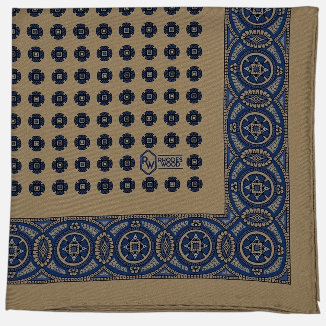 A camel 100% silk pocket square with navy blue medallions repeating across the silk .