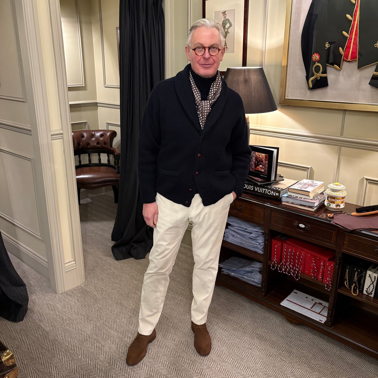 Man in a stylish outfit standing in an elegant room with books and a lamp.