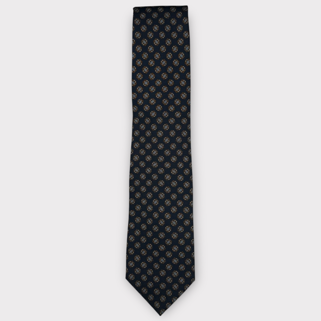 A classic necktie with a repeating geometric medallion pattern in dark navy and gold, displayed flat against a light gray background.