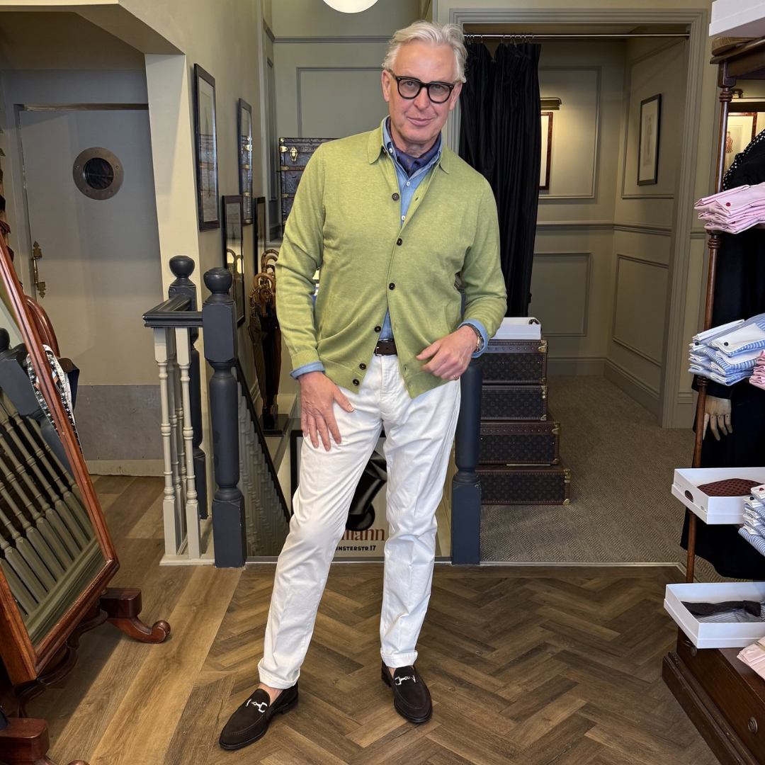 Man in a green cardigan and white pants standing in a store with suitcases and clothing racks.