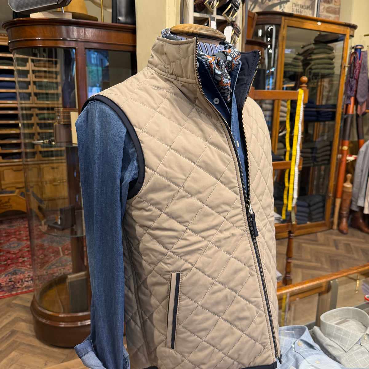 A men's quilted zip-front vest in light beige/tan displayed on a mannequin torso, layered over a blue shirt and patterned scarf, inside a vintage clothing store with wooden display cases.