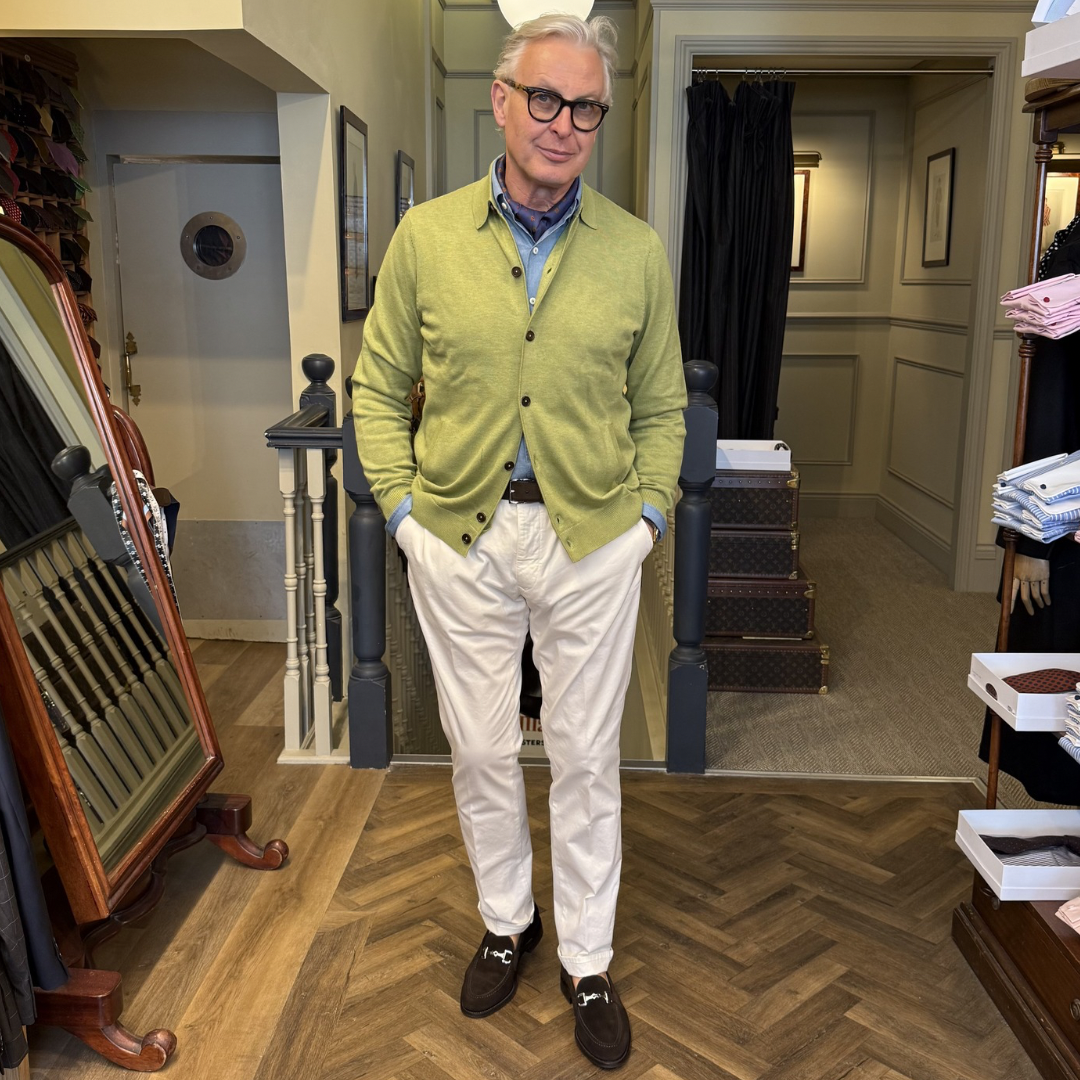 Man wearing a green cardigan and white pants standing in a store with suitcases and clothing racks.